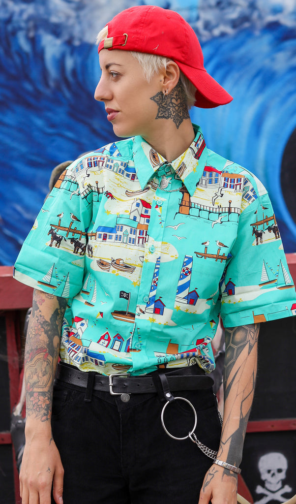 Model is stood in front of a wave mural wearing the Brighton seaside short sleeve shirt with a red cap and black jeans. They are facing the camera and looking to the left with both hands by their sides. They are heavily tattooed and have short white blonde hair. The photo is cropped from the hips up.