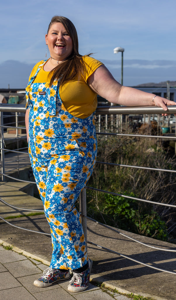 Nat is stood outside leaning on a metal railing wearing the blue and yellow floral cotton twill dungarees with a short sleeved yellow tshirt with multicoloured trainers. They are facing forward leaning back with one hand up on the railing and the other in their dungaree pocket whilst laughing to camera.