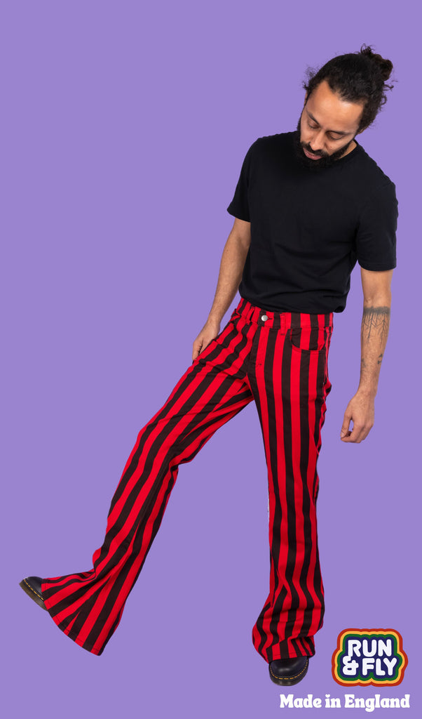 Richard, a hispanic male model with dark hair in a bun and a beard, is stood in a photography studio in Hove in front of a lilac backdrop wearing Red & Black Striped Bell Bottom Stretch Super Flares with a black t shirt and black shoes. Richard is facing the camera with one leg turned out to the side and lifted in the air. He is looking down with his arms down by his sides. Run & Fly logo is at the corner or the photo with 'Made in England' written underneath.