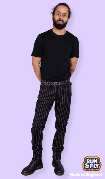 Richard, a hispanic male model with dark hair in a bun and a beard, is stood in a photography studio in front of a lilac backdrop wearing Stretch Black & White Pinstriped Skinny Jeans with a black t shirt tucked in, a checkered belt and black boots. He is facing the camera with one leg slightly bent and his arms behind his back. Run & Fly logo is at the corner or the photo with 'Made in England' written underneath.