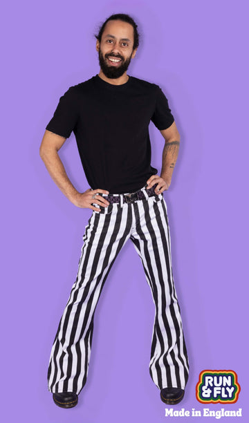 Richard, a hispanic male model with dark hair in a bun and a beard, is stood in a photography studio in front of a lilac backdrop wearing Black & White Striped Bell Bottom Stretch Super Flares with a black t shirt, black and purple checkered belt and black shoes. Richard is smiling and facing the camera with his hands on his hips. Run & Fly logo is at the corner or the photo with 'Made in England' written underneath.