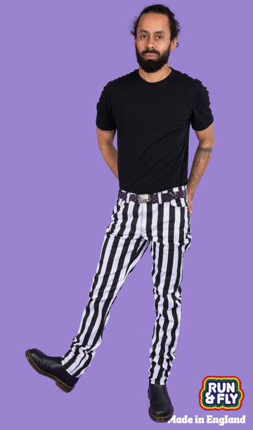 Richard, a hispanic male model with dark hair in a bun and a beard, is stood in a photography studio in Hove in front of a lilac backdrop wearing Black & White 1" Striped Mid Rise Stretch Skinny Jeans with a purple and black checkered belt, a black t shirt and black shoes. Richard is posing facing the camera with one leg turned out to the side and arms behind their back. Run & Fly logo is at the corner or the photo with 'Made in England' written underneath.