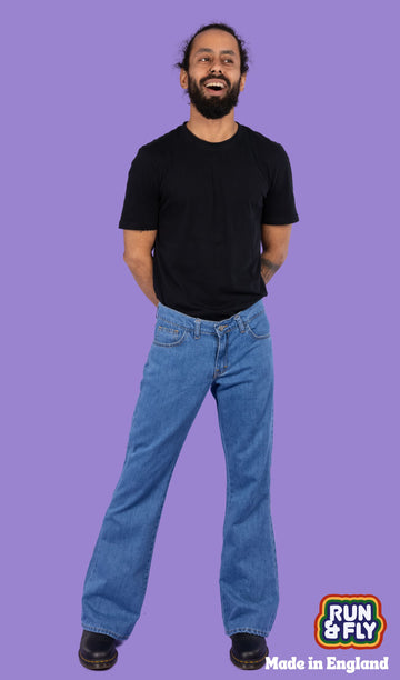 Richard, a hispanic male model with dark hair in a bun and a beard, is stood in a photography studio in Hove in front of a lilac backdrop wearing Stonewash Retro Boot Cut Flare Jeans with a black t shirt and black shoes. Richard is smiling and facing the camera with his arms behind his back and legs slightly apart. Run & Fly logo is at the corner or the photo with 'Made in England' written underneath.