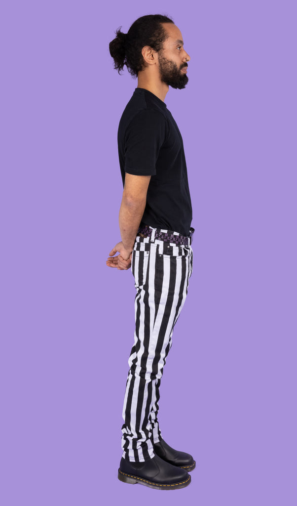 Richard, a hispanic male model with dark hair in a bun and a beard, is stood in a photography studio in Hove in front of a lilac backdrop wearing Black & White 1" Striped Mid Rise Stretch Skinny Jeans with a purple and black checkered belt, a black t shirt and black shoes. Richard is posing sideways with his arms behind his back. 