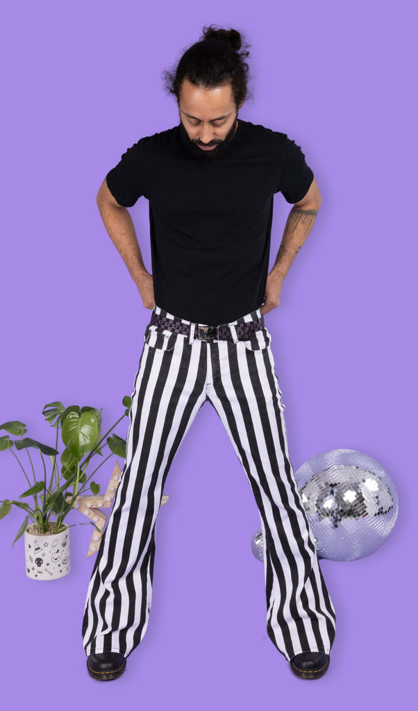 Richard, a hispanic male model with dark hair in a bun and a beard, is stood in a photography studio in front of a lilac backdrop amongst disco balls, a plant in a pot and a star shaped light wearing Black & White Striped Bell Bottom Stretch Super Flares with a black t shirt, black and purple checkered belt and black shoes. Richard is facing the camera and is looking down pulling up trousers with both hands.