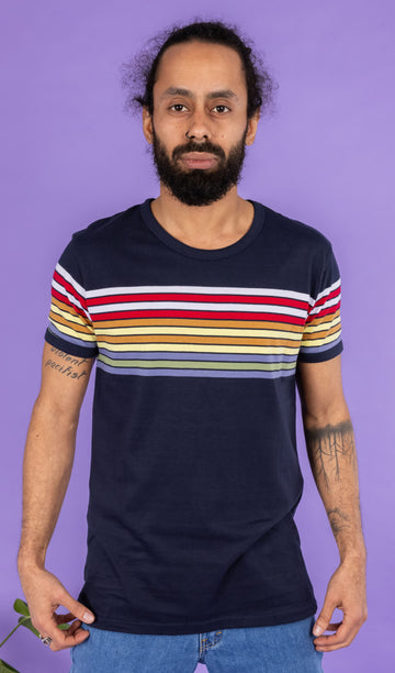 Richard, a hispanic male model with dark hair in a bun and a beard, is stood in a photography studio in Hove in front of a lilac backdrop wearing Navy Stripe Tee with blue jeans. Richard is posing toward the camera, slightly pulling down the t shirt with his hands. The photo is cropped at the thighs.