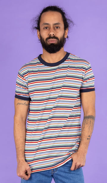 Richard, a hispanic male model with dark hair in a bun and a beard, is stood in a photography studio in Hove in front of a lilac backdrop wearing Retro Multi Striped Ringer Tee with blue jeans. The short sleeved t shirt has horizontal stripes going across it in navy blue, white, orange, brown, yellow, pastel blue and green. Richard is posing facing the camera and is pulling down the bottom of the t shirt. The photo is cropped at the thighs.