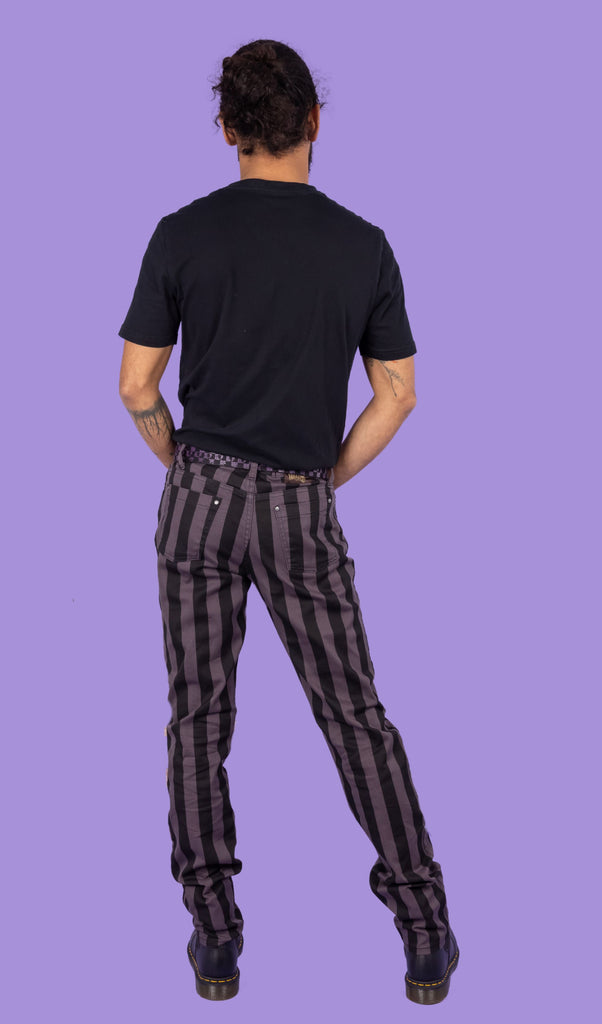 Richard, a hispanic male model with dark hair in a bun and a beard, is stood in a photography studio in Hove in front of a lilac backdrop wearing Black & Grey 1" Striped Mid Rise Stretch Skinny Jeans with a purple and black checkered belt, a black t shirt and black shoes. Richard is posing with his back to the camera and is leaning to one side with his arms in front of him.