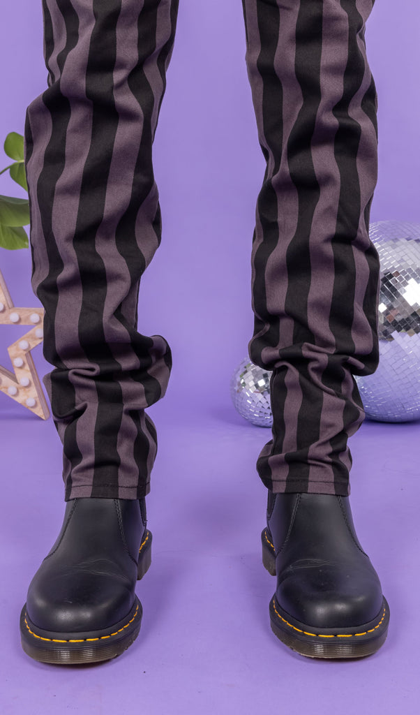 Close up of Richard wearing the Black & Grey 1" Striped Mid Rise Stretch Skinny Jeans with black shoes. The background of the photo is lilac and their are disco balls behind him. The photo is cropped at the knees.