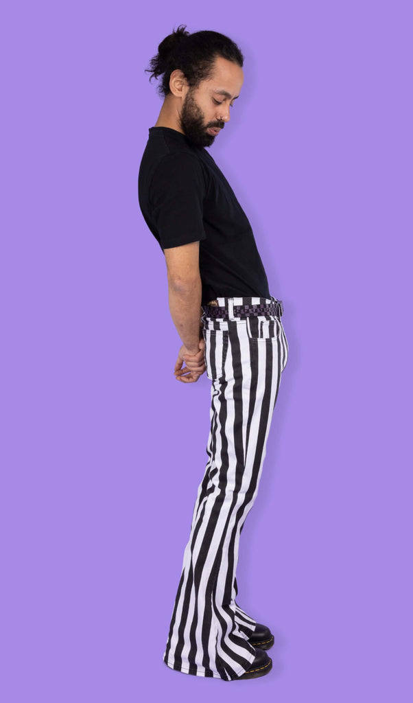 Richard, a hispanic male model with dark hair in a bun and a beard, is stood in a photography studio in front of a lilac backdrop wearing Black & White Striped Bell Bottom Stretch Super Flares with a black t shirt, black and purple checkered belt and black shoes. Richard is stood sideways looking down and bending back slightly with his arms behind his back. 