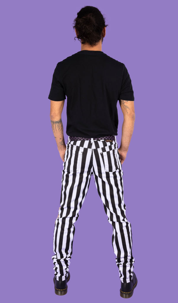 Richard, a hispanic male model with dark hair in a bun and a beard, is stood in a photography studio in Hove in front of a lilac backdrop wearing Black & White 1" Striped Mid Rise Stretch Skinny Jeans with a purple and black checkered belt, a black t shirt and black shoes. Richard is posing with his back to the camera and thumbs in the front pockets of the jeans.
