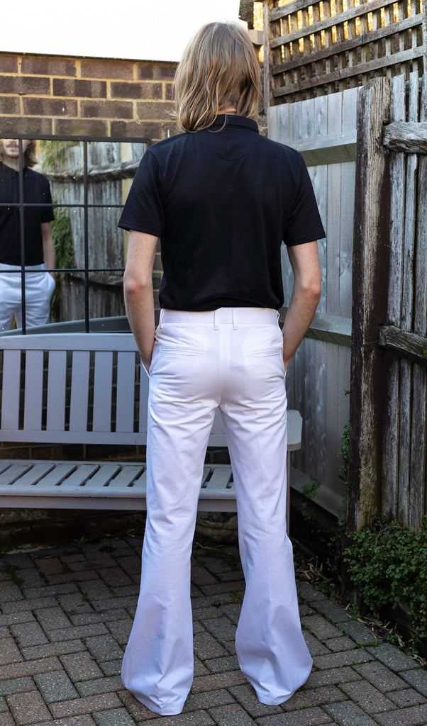 Jack is stood outside in a garden area wearing the Presley white cotton bell bottom trouser slacks with a short sleeve black shirt and white trainers. He is facing away from the camera with his hands in the pockets of the trousers.