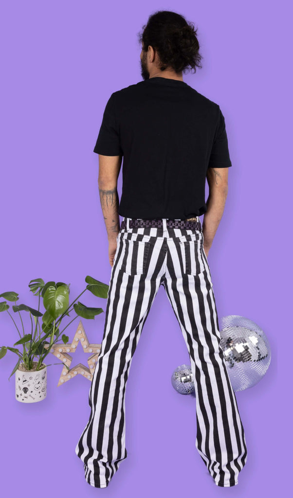 Richard, a hispanic male model with dark hair in a bun and a beard, is stood in a photography studio in front of a lilac backdrop amongst disco balls, a plant in a pot and a star shaped light wearing Black & White Striped Bell Bottom Stretch Super Flares with a black t shirt, black and purple checkered belt and black shoes. Richard has his back to the camera and his arms down by his sides.