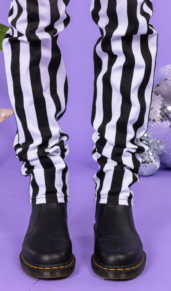 Close up of Richard wearing the Black & White 1" Striped Mid Rise Stretch Skinny Jeans with black shoes. The background of the photo is lilac and their are disco balls behind him. The photo is cropped at the knees. 