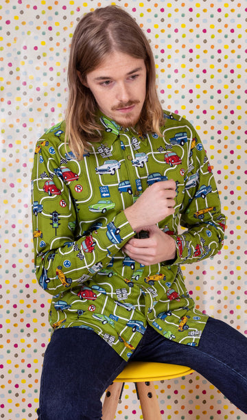 Jack is sat on a yellow stool in front of a multicoloured polka dot background wearing the race track car play button down long sleeve shirt with denim jeans. They are facing forward and posing doing up the sleeve cuff buttons whilst looking down to the right. Photo is cropped from the hips up.