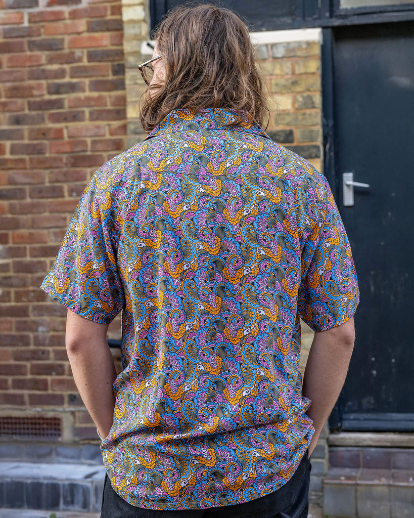 Michael is a white male and has shoulder length sandy hair, he is wearing a pink, blue and gold paisley print 70's style relaxed shirt and standing in a bricked mews he has his back to the camera and is looking off to the side.