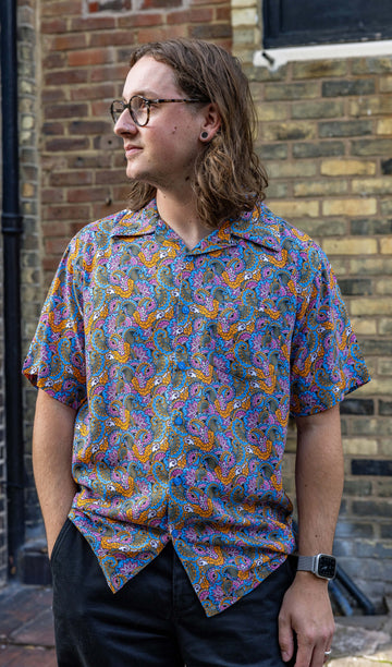 Michael is a white male and has shoulder length sandy hair, nose ring and glasses he is wearing a  pink, blue and gold paisley print paisley 70's style relaxed shirt and standing in a bricked mews looking away from camera. 