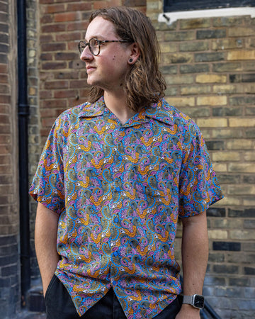 Michael is a white male and has shoulder length sandy hair, nose ring and glasses he is wearing a  pink, blue and gold paisley print paisley 70's style relaxed shirt and standing in a bricked mews looking away from camera. 