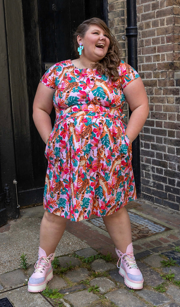 Natalie is wearing Tiger Lily Stretch Belted Tea Dress with Pockets with cactus shaped earrings and pink boots. The baby pink dress has an all over print of tigers with flowers and leaves. Natalie is stood outside in Hove facing the camera with her hands in the dress pockets and is smiling looking off to one side.