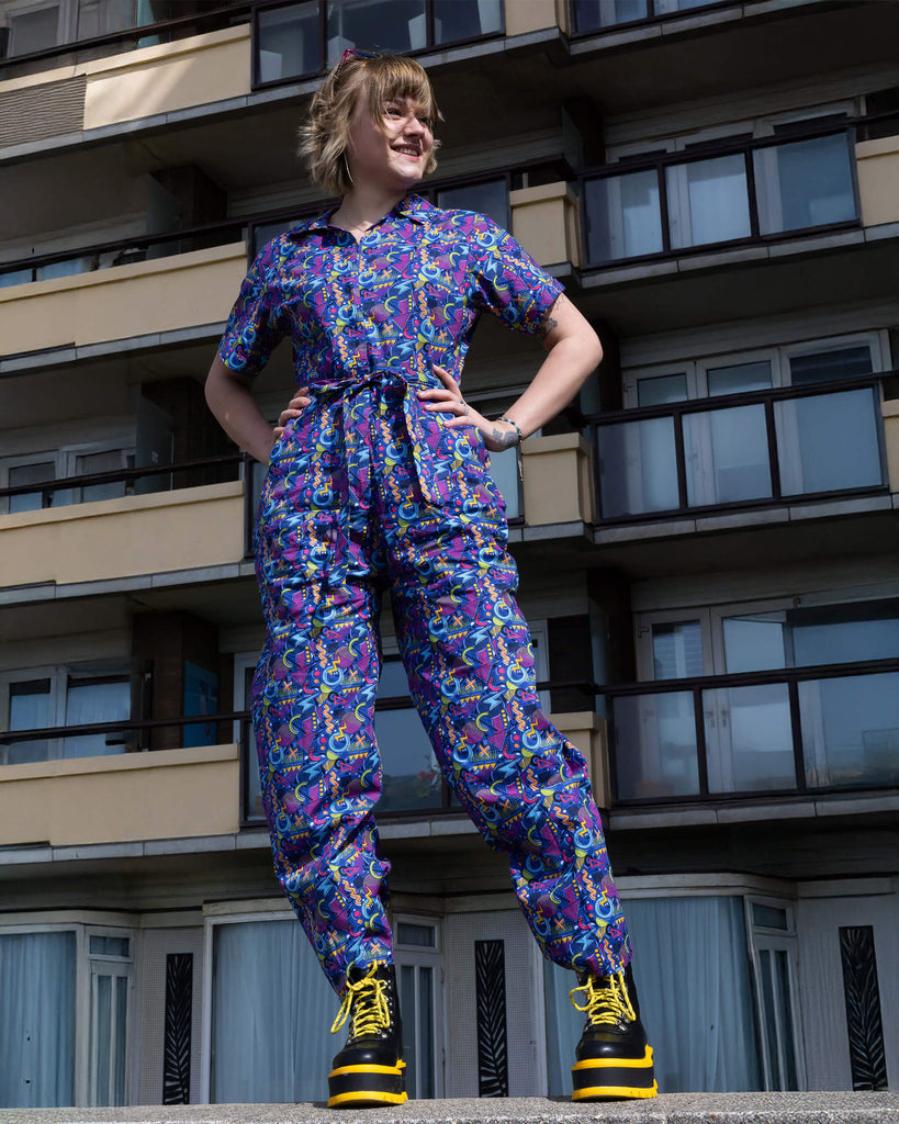 The 90's Arcade Jumpsuit being worn by a femme model with short blonde hair with koi footwear yellow and black platform boots. She is stood posing with both hands on her hips looking off to the right standing on top of a concrete wall in front of a block of flats. The jumpsuit print features classic 90's style shapes, squiggles and doodles in green, orange, pink, purple and light blue all on a dark blue background.