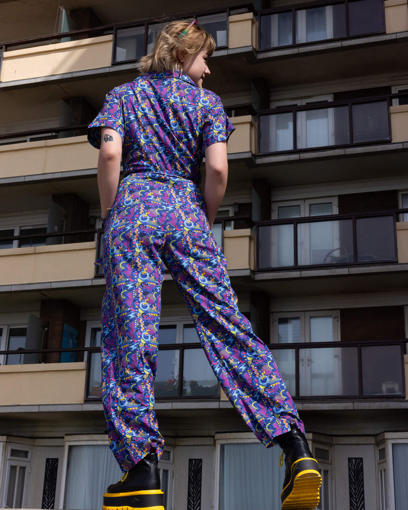 The 90's Arcade Jumpsuit being worn by a femme model with short blonde hair with koi footwear yellow and black platform boots. She is stood posing away from camera with both hands in the side pockets looking off to the right standing on top of a concrete wall in front of a block of flats. The jumpsuit print features classic 90's style shapes, squiggles and doodles in green, orange, pink, purple and light blue all on a dark blue background.
