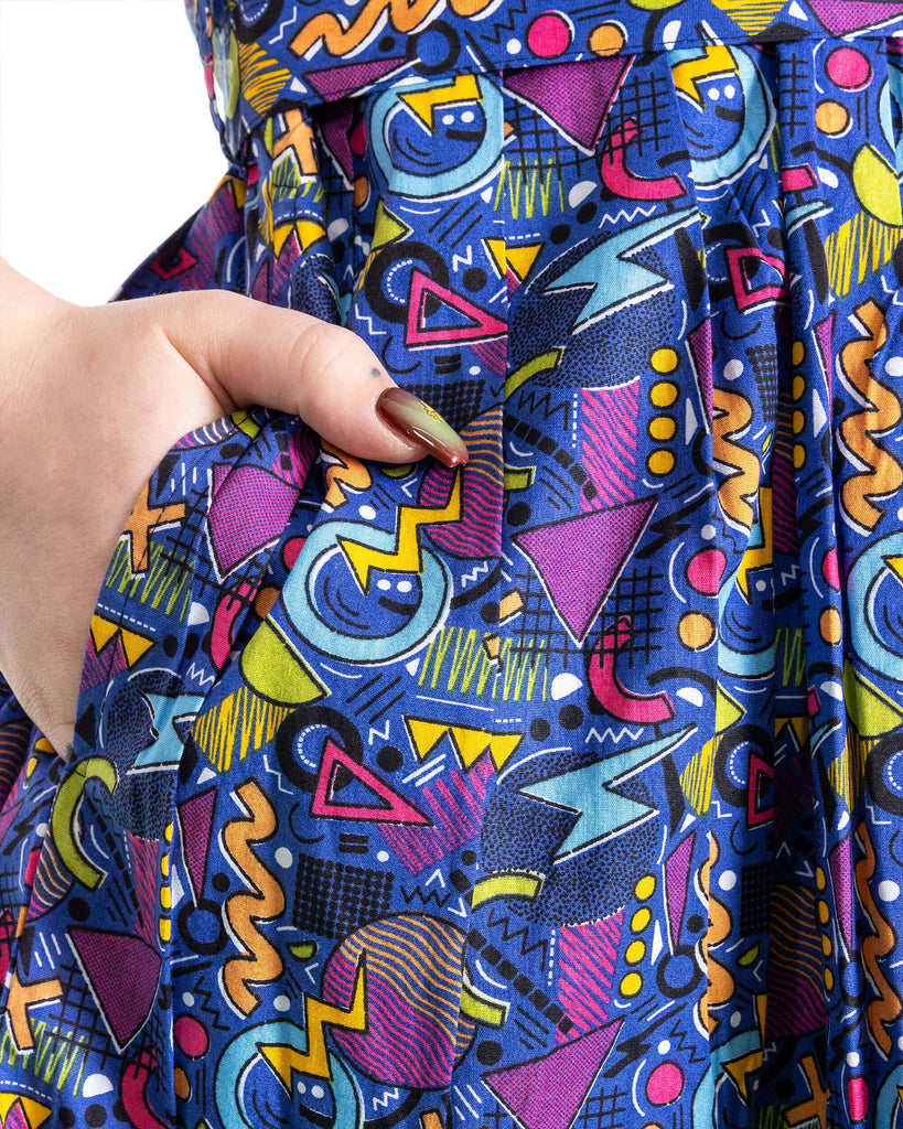 Close up of model's hand in the pocket of the 90's Arcade Stretch Belted Tea Dress with Pockets. The print features classic 90's style shapes, squiggles and doodles in green, orange, pink, purple and light blue all on a dark blue background.
