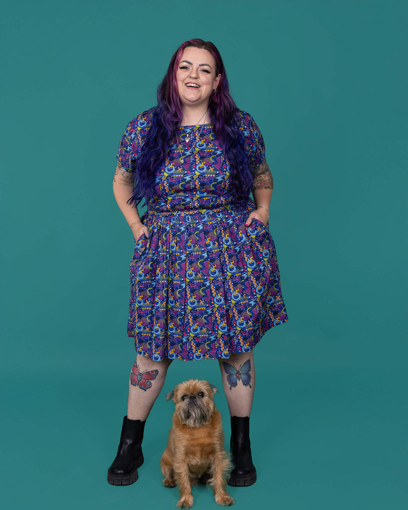 The 90's Arcade Stretch Belted Tea Dress with Pockets worn by a femme alternative tattooed model with long pink to purple hair and black boots on a teal studio background with a small brown dog. She is facing forward laughing with both hands in the dress pockets. The print features classic 90's style shapes, squiggles and doodles in green, orange, pink, purple and light blue all on a dark blue background.