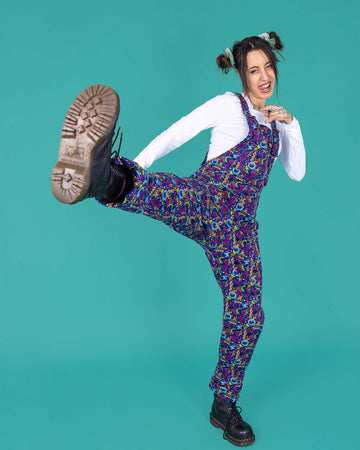 Yo is a femme model with bunches and doing a high kick to camera. They are wearing 90's inspired carpet dungarees in blues, pink and orange.