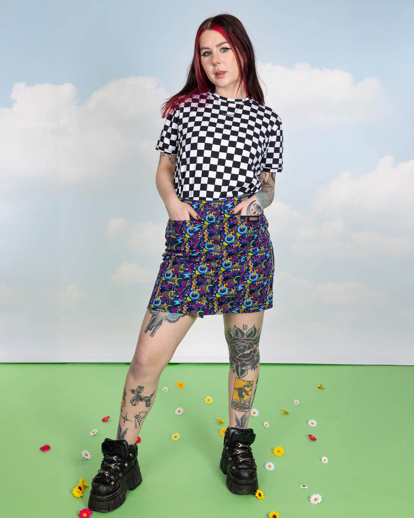 Florence is stood in a photography studio in Hove in front of blue sky backdrop on a green floor with flowers scattered wearing 90's Arcade A Line Skirt with black and white checker board short sleeve tee and black shoes. The skirt print features classic 90's style shapes, squiggles and doodles in green, orange, pink, purple and light blue all on a dark blue background. Florence is posing with her hands in the skirt pockets.