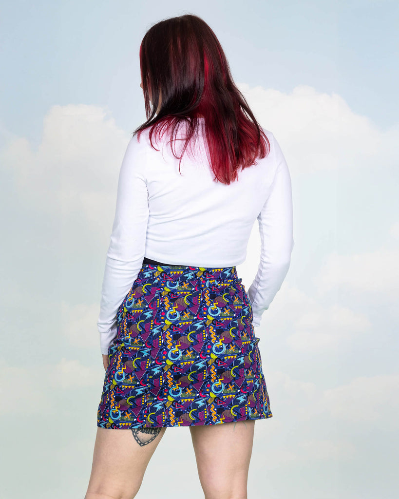 Florence is stood in a photography studio in Hove in front of blue sky backdrop wearing 90's Arcade A Line Skirt with a long sleeved white top. The skirt print features classic 90's style shapes, squiggles and doodles in green, orange, pink, purple and light blue all on a dark blue background. Florence is posing with her back to the camera and hands resting by her sides.