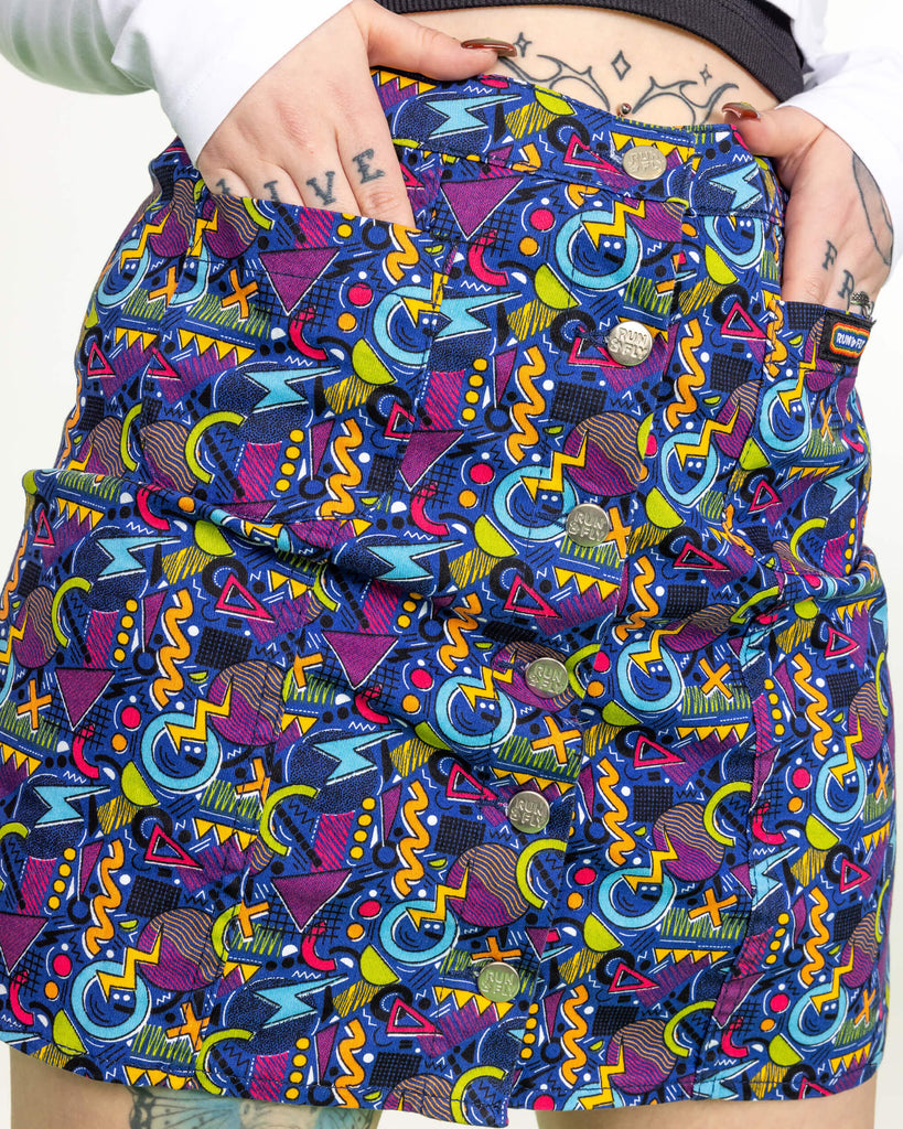 Close up model's hands in the pockets of the 90's Arcade A Line Skirt. The skirt print features classic 90's style shapes, squiggles and doodles in green, orange, pink, purple and light blue all on a dark blue background.