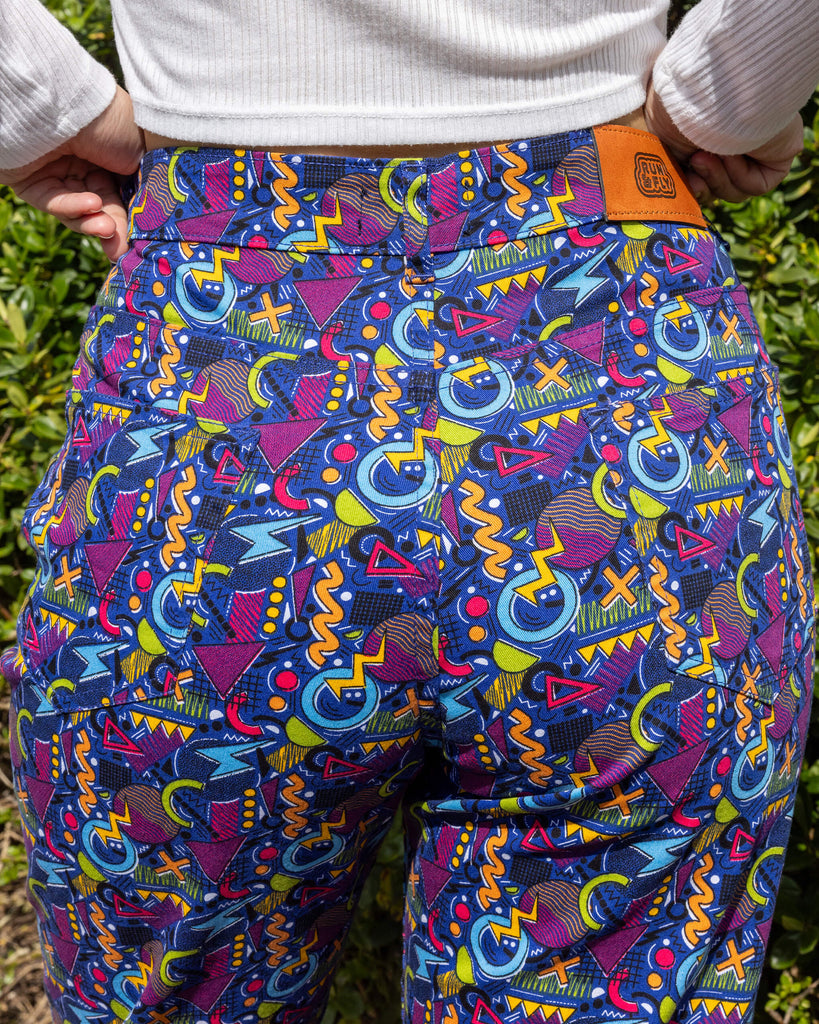 Close up in model's hand in the back pocket of the 90's Arcade High Waist Straight Leg Jeans. The jeans print features classic 90's style shapes, squiggles and doodles in green, orange, pink, purple and light blue all on a dark blue background. Close up shows the Run & Fly logo on the back.
