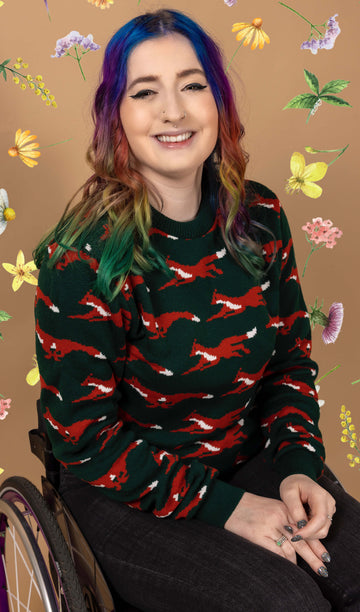 Eliza, a femme model with rainbow coloured hair, is sat in their wheelchair in front of a tan backdrop with flowers on it. They are wearing the Running Fox Jumper and smiling at the camera. 