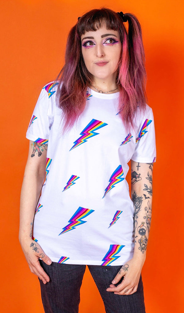 Sarah is stood wearing the white lightning bolt short sleeve tee with black stonewash denim flares. She has pink and brown hair in bunches with pink makeup and arm tattoos. She is facing towards the camera with both arms resting by her side and smiling looking up to the right. The tshirt is a white base colour with repeating rainbow lightning bolts varying in size all over.