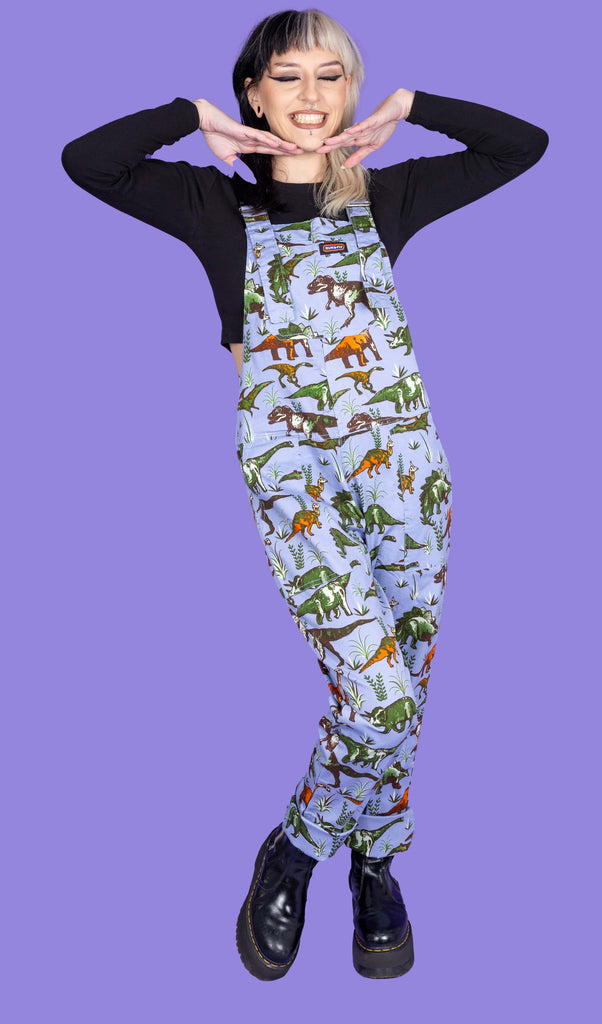 Model with split dyed black and blonde hair is wearing Blue Adventure Dinosaur Stretch Twill Dungarees paired with a long sleeve black top underneath and black boots. The dungarees are a baby blue colour with various dinosaurs in orange and green. The model is smiling with hands under her chin.