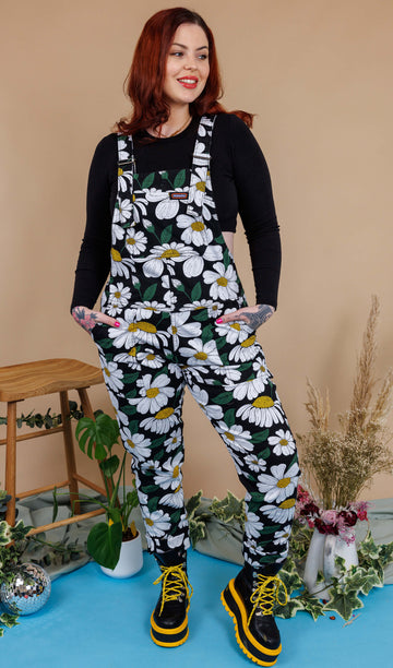 A red haired tattooed female white model smiling wearing Daisy Dungarees and black crop top against a beige background with flowers, a stool and mirror ball
