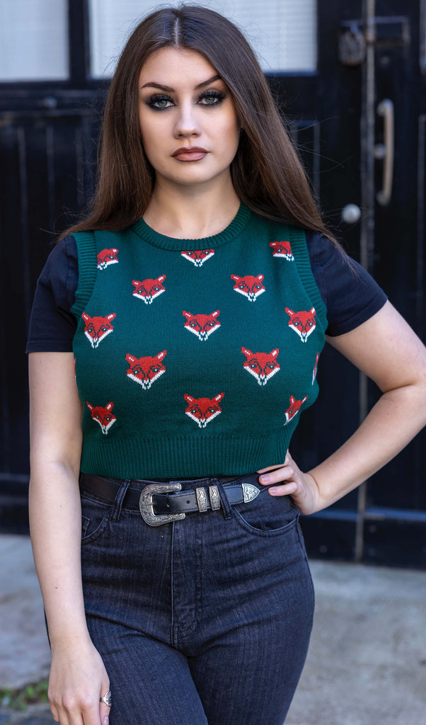 Charlotte is stood outside in a courtyard wearing the fox head green knitted tank top with a short sleeve black tshirt underneath and black stone wash flares. She is facing forward with one hand on her hip whilst looking to camera. The knitted tank top is a bottle green colour with various orange and white fox heads all over and a plain green back.