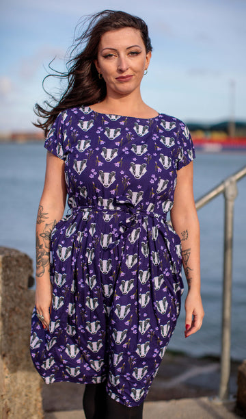 a white femme model wearing Badger Tea Party Dress by Run & Fly. The blue dress has an all over badger head print with purple flowers amongst them. There is a matching fabric belt tied around the model's waist. The model is stood in front of a river posing. 