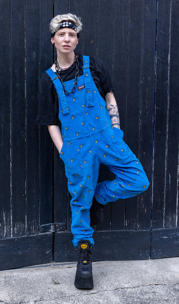 Jamie a white Masc trans man with blonde short hair is wearing blue corduroy bee printed dungarees, black boots and a black tee with big black chains. He is leaning on a black door in a mews in Hove shot by Amy Davies Photography