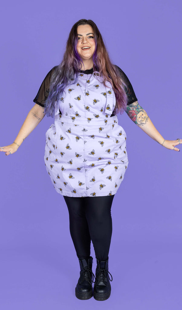 Luisa stood in a photography studio in Hove in front of a purple backdrop wearing Lavender Bees Stretch Twill Pinafore Dress with a short sleeve mesh top underneath, black tights and boots. The lavender pinafore has an all over bumble bee print. Luisa is smiling facing the camera with her arms out by her sides.