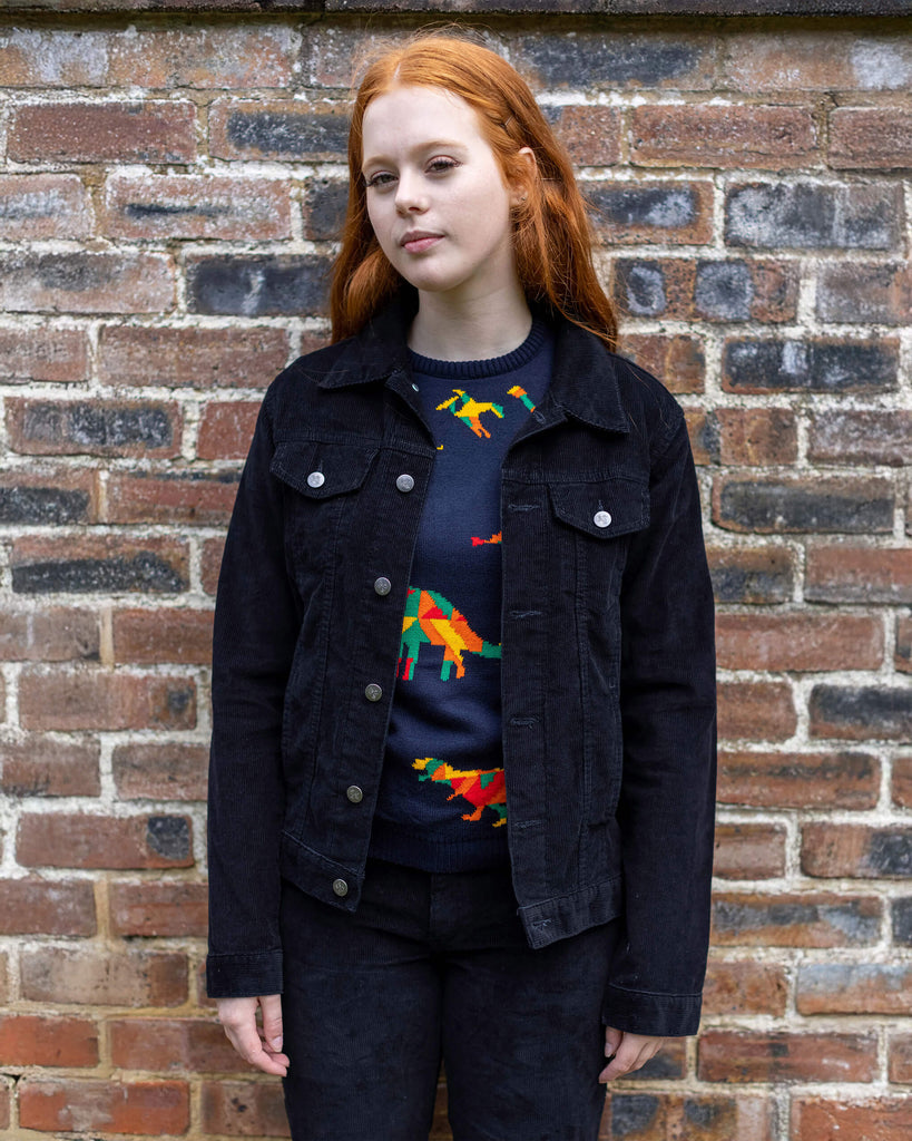 Sophie is stood in front of a brick wall wearing the retro vintage black cord unisex western jacket with the rainbow dino jumper and black trousers. She is facing forward with both arms resting by her sides. Photo is cropped from the knees up.
