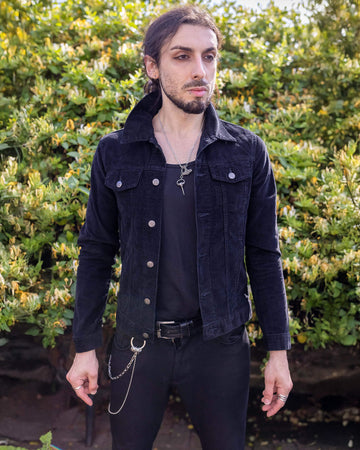 Riccardo is stood outside in a garden area wearing the retro vintage black cord unisex western jacket with black trousers. They are facing away from the camera to highlight the back waist adjustable tabs. They are facing towards the camera with both hands resting by their side. Photo is cropped from the knees up.