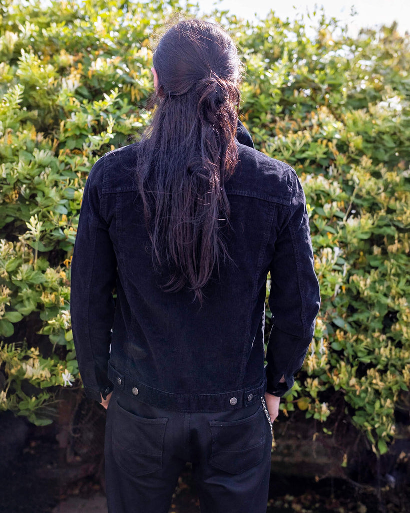 Riccardo is stood outside in a garden area wearing the retro vintage black cord unisex western jacket with black trousers. They are facing away from the camera to highlight the back waist adjustable tabs. They are facing away from the camera with both hands in their jean pockets. Photo is cropped from the knees up.