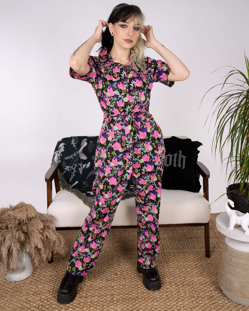 The model adjusts their hair while wearing the Run & Fly x Marie Boiseau Bodies Stretch Jumpsuit. Featuring a bold, abstract print of bodies and plants, the black jumpsuit showcases sustainable style. Decor elements like gothic cushions and pampas grass complete the look.