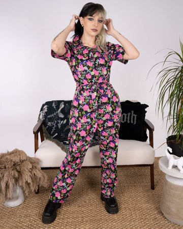 The model adjusts their hair while wearing the Run & Fly x Marie Boiseau Bodies Stretch Jumpsuit. Featuring a bold, abstract print of bodies and plants, the black jumpsuit showcases sustainable style. Decor elements like gothic cushions and pampas grass complete the look.