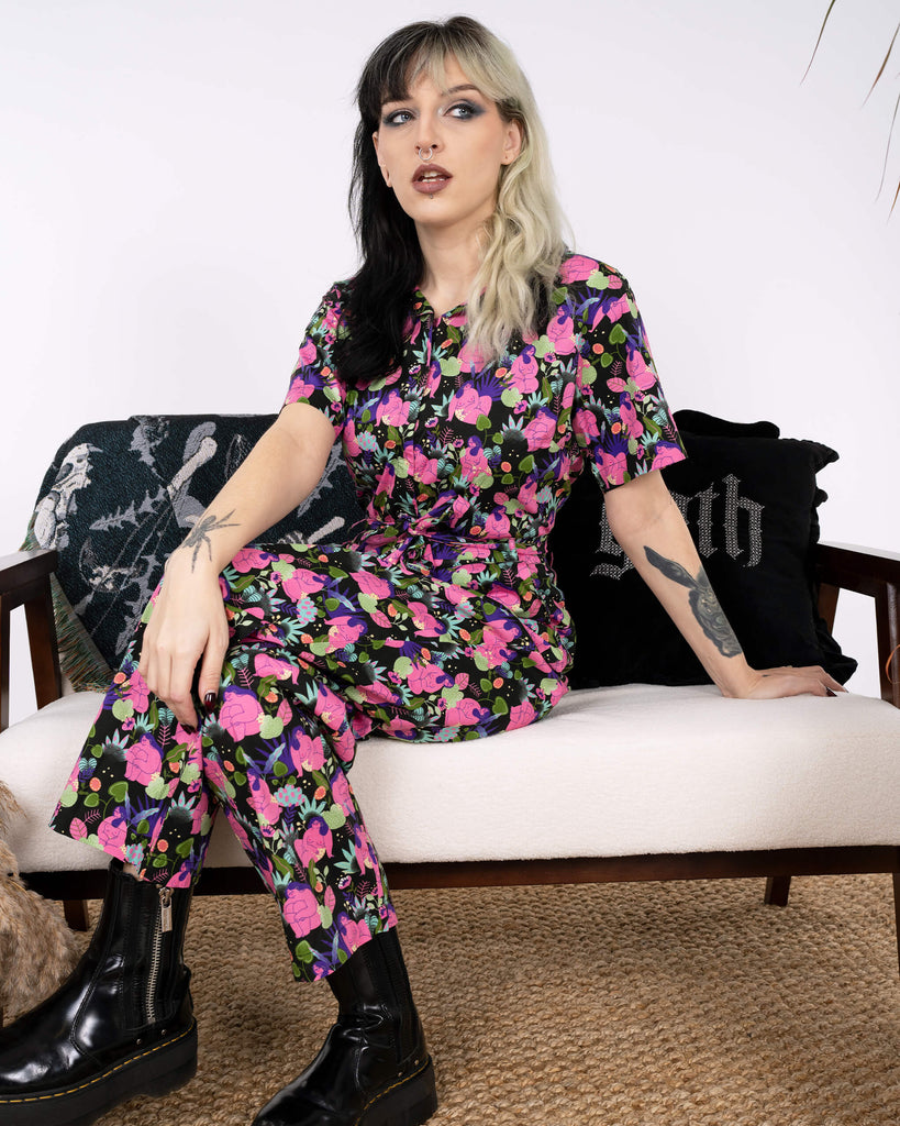 The model sits stylishly on a white bench wearing the Run & Fly x Marie Boiseau Bodies Stretch Jumpsuit. The bold black jumpsuit with artistic pink and purple body patterns exudes creativity. Ethical slow fashion meets comfort and statement style. A gothic-themed cushion and pampas grass add charm.