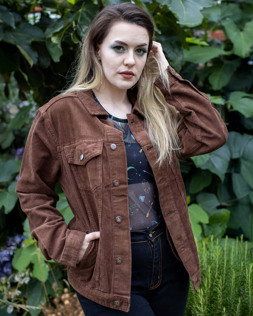 Alice is stood outside in a garden area wearing the retro vintage cocoa brown cord unisex western jacket with a black tshirt and black jeans. She is facing the camera posing with one hand in her jacket pocket whilst the other hand is pushing back her hair whilst looking to camera. Photo is cropped from the hips up.