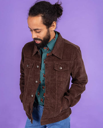 Richard, a hispanic male model with dark hair in a bun and a beard, is stood in a photography studio in Hove in front of a lilac backdrop wearing Retro Vintage Cocoa Brown Corduroy Unisex Western Jacket with Preppy Foxy Short Sleeve Shirt underneath and blue jeans. Richard is posing to one side and is looking down with his hands in the pockets of the jacket. The photo is cropped at the thigh.