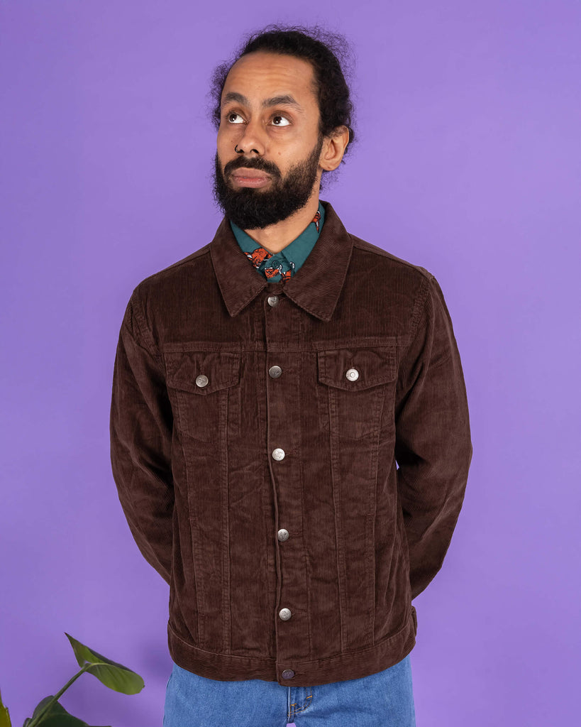 Richard, a hispanic male model with dark hair in a bun and a beard, is stood in a photography studio in Hove in front of a lilac backdrop wearing Retro Vintage Cocoa Brown Corduroy Unisex Western Jacket buttoned up with Preppy Foxy Short Sleeve Shirt underneath and blue jeans. Richard is posing facing the camera and looking up to one side with his arms behind his back. The photo is cropped at the thighs.