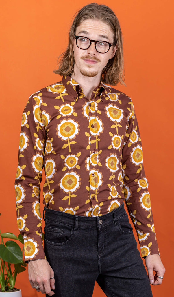 Jack a white male with shoulder length blonde hair and glasses in his 20's wearing a retro brown shirt with long selves and black jeans shot against an orange studio background